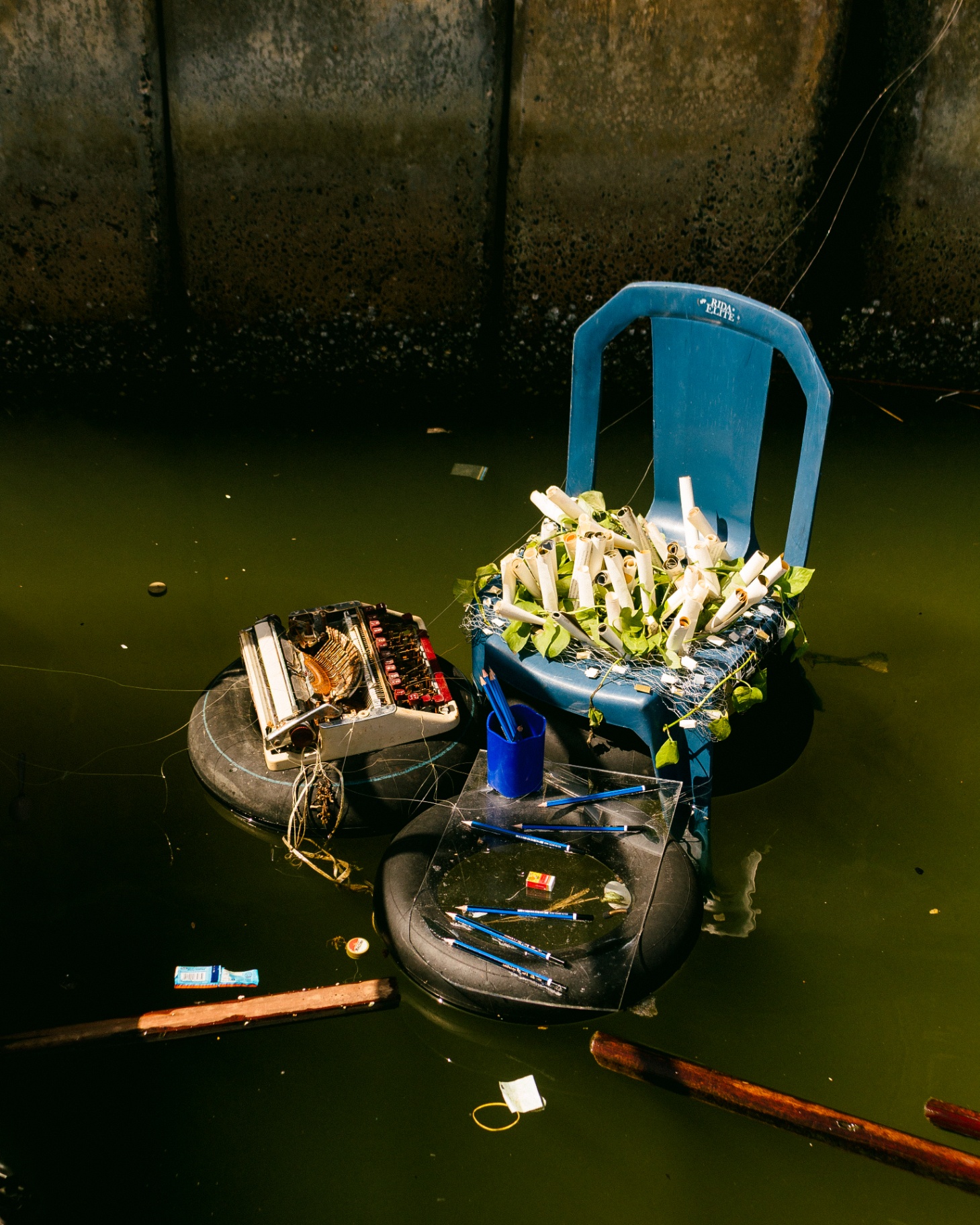A floating installation of a blue plastic chair holding rolled papers, a rusted typewriter and pencils drifting on the surface of the Lagos Lagoon