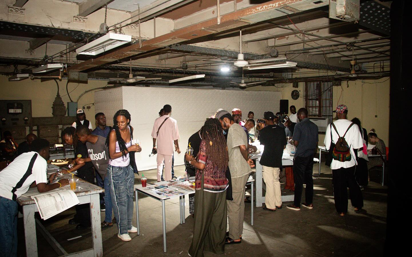 Wide view of participants moving between print and collage tables inside the exposed-beam hall of the former Government Printing Press at Kedu Lagos