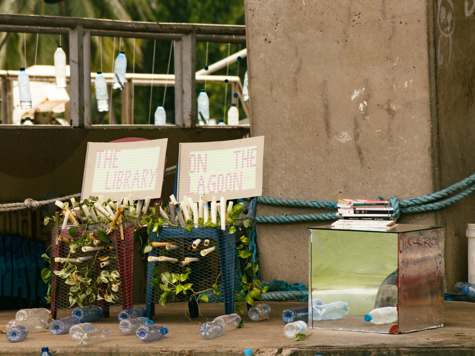 Wire-mesh stools stuffed with paper scrolls and pothos vines beside a mirrored cube, with a spiral-bound book propped on top and plastic water bottles scattered on the ground