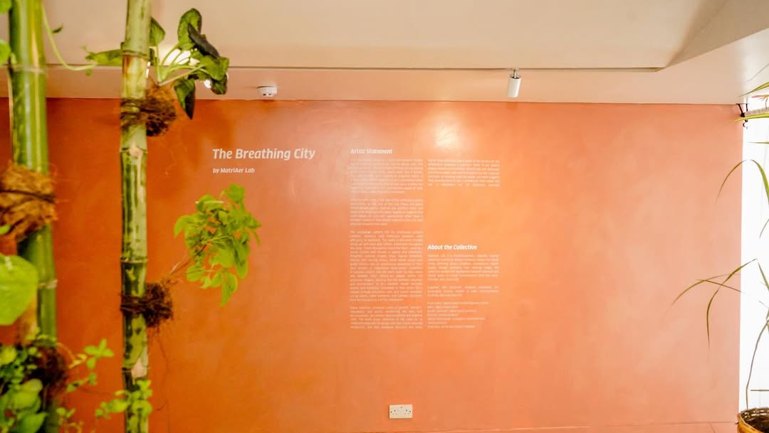 The Breathing City wall text by MatriAér Lab painted on a terracotta wall with hanging bamboo and plants