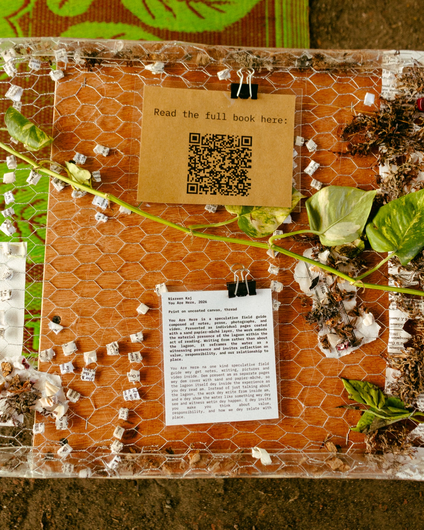 Top-down view of a research plinth wrapped in chicken wire, a QR code, a printed text panel by Nisreen Kaj, and pothos leaves arranged over paper fragments