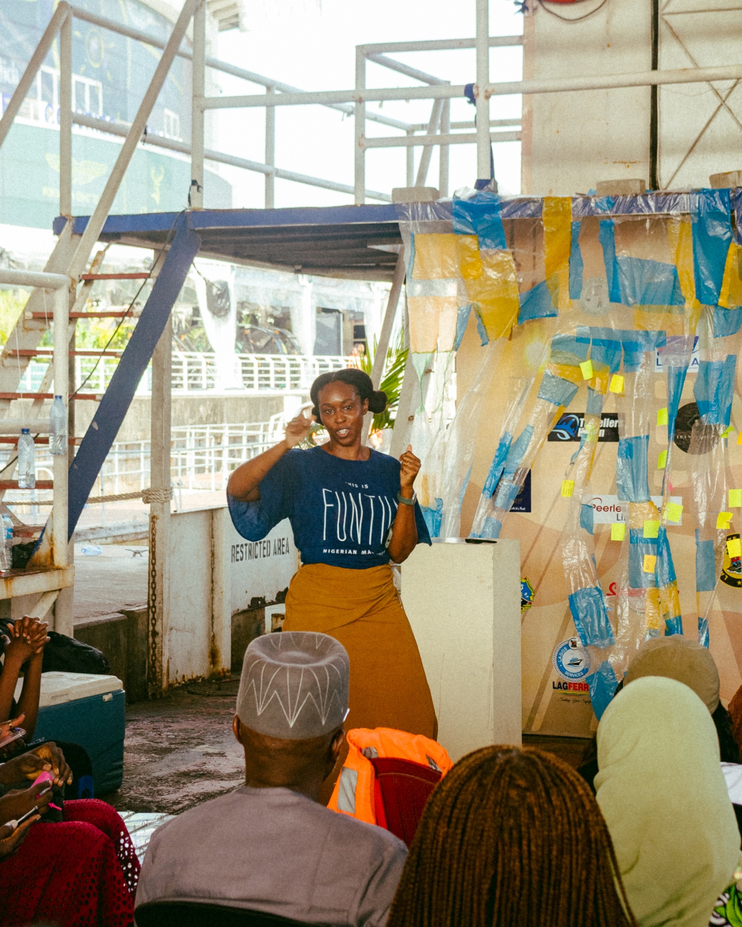 A speaker in a 'This Is Funtime Nigerian Made' t-shirt addresses the audience beside a plastic-woven installation of tape, receipts and stickers at Library on the Lagoon