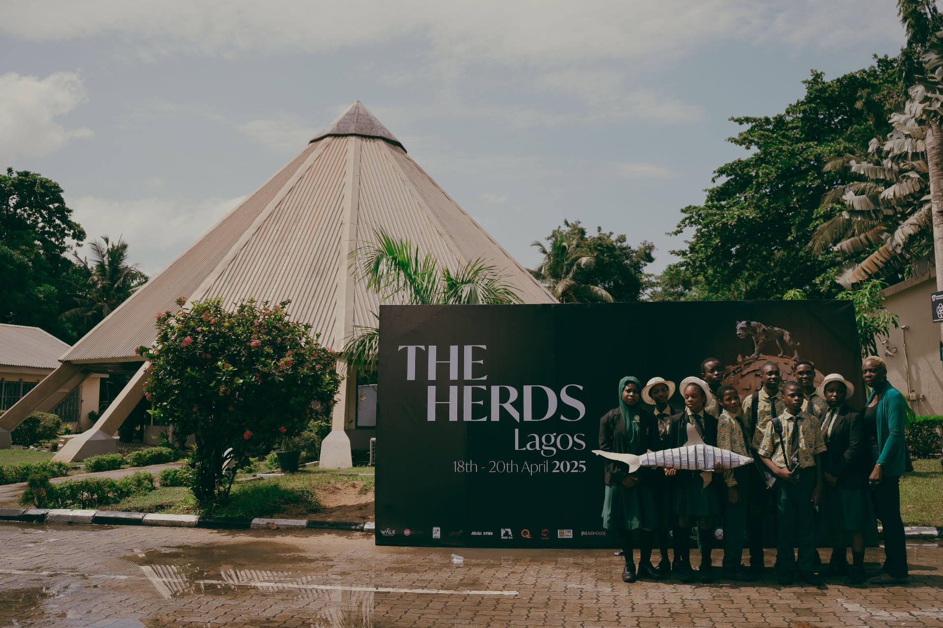 The Herds Lagos signage outside the venue with schoolchildren holding a fish sculpture