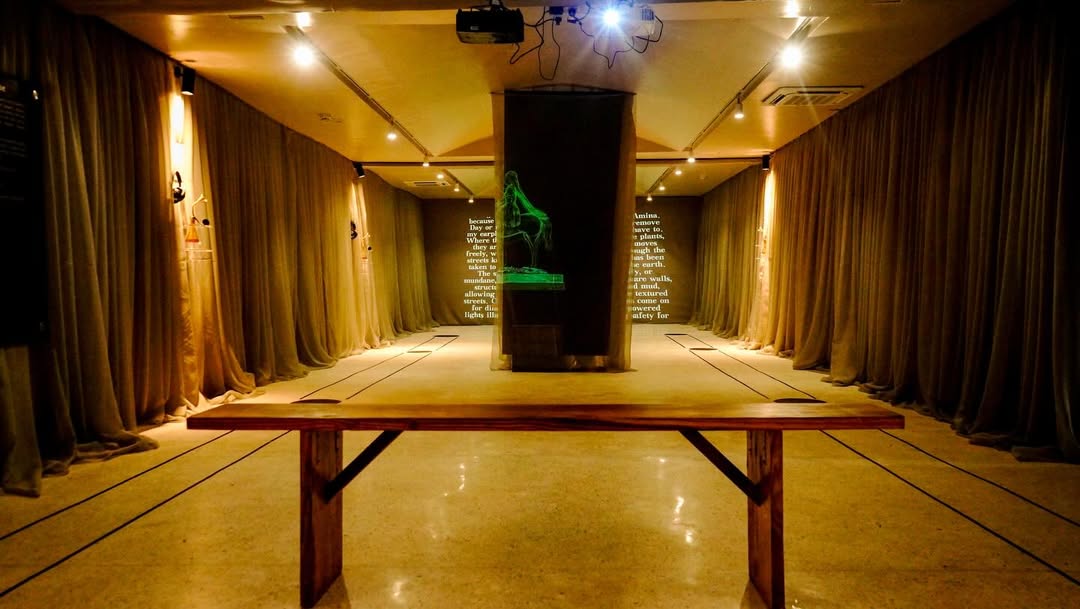 Central plinth with a glowing green Queen Amina figurine in the Sarauniya Ènè installation, framed by curtained walls and a long wooden bench