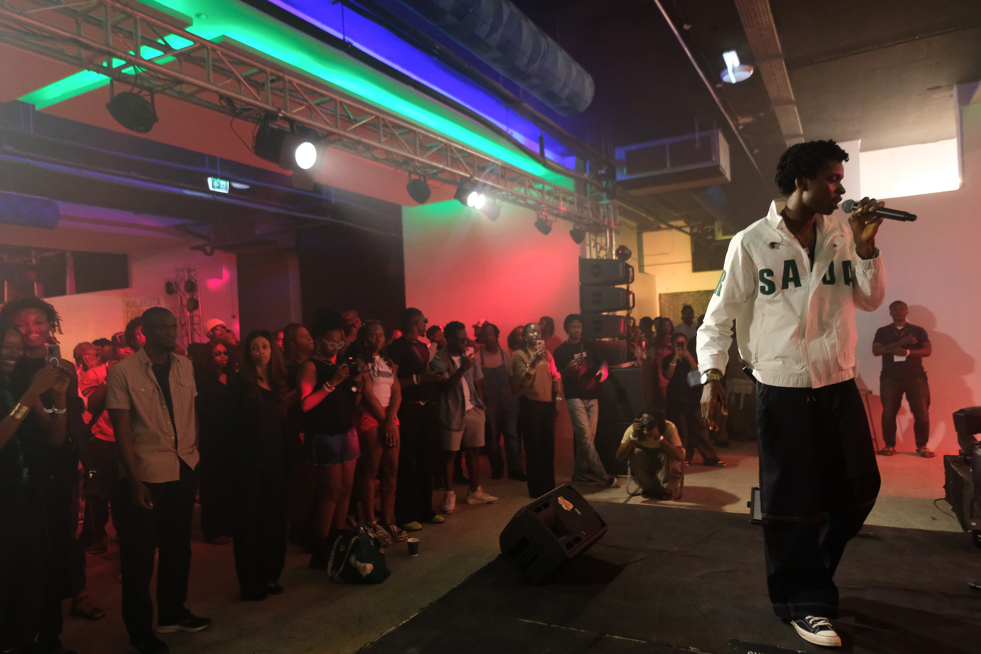 MC in a white Saba jacket addressing the crowd under green and red stage lights at Afrobeat Rebellion