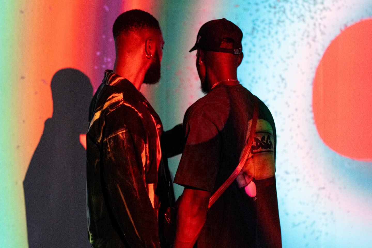 Two visitors silhouetted in red and blue projected light in front of a speckled digital artwork at Kedu Lagos