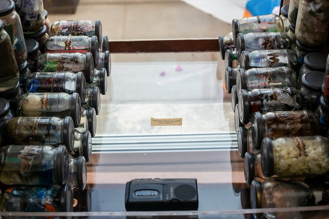 Overhead view of jars containing artefacts flanking a printed label reading 'Should Lagos survive the water?' with a small cassette recorder between them