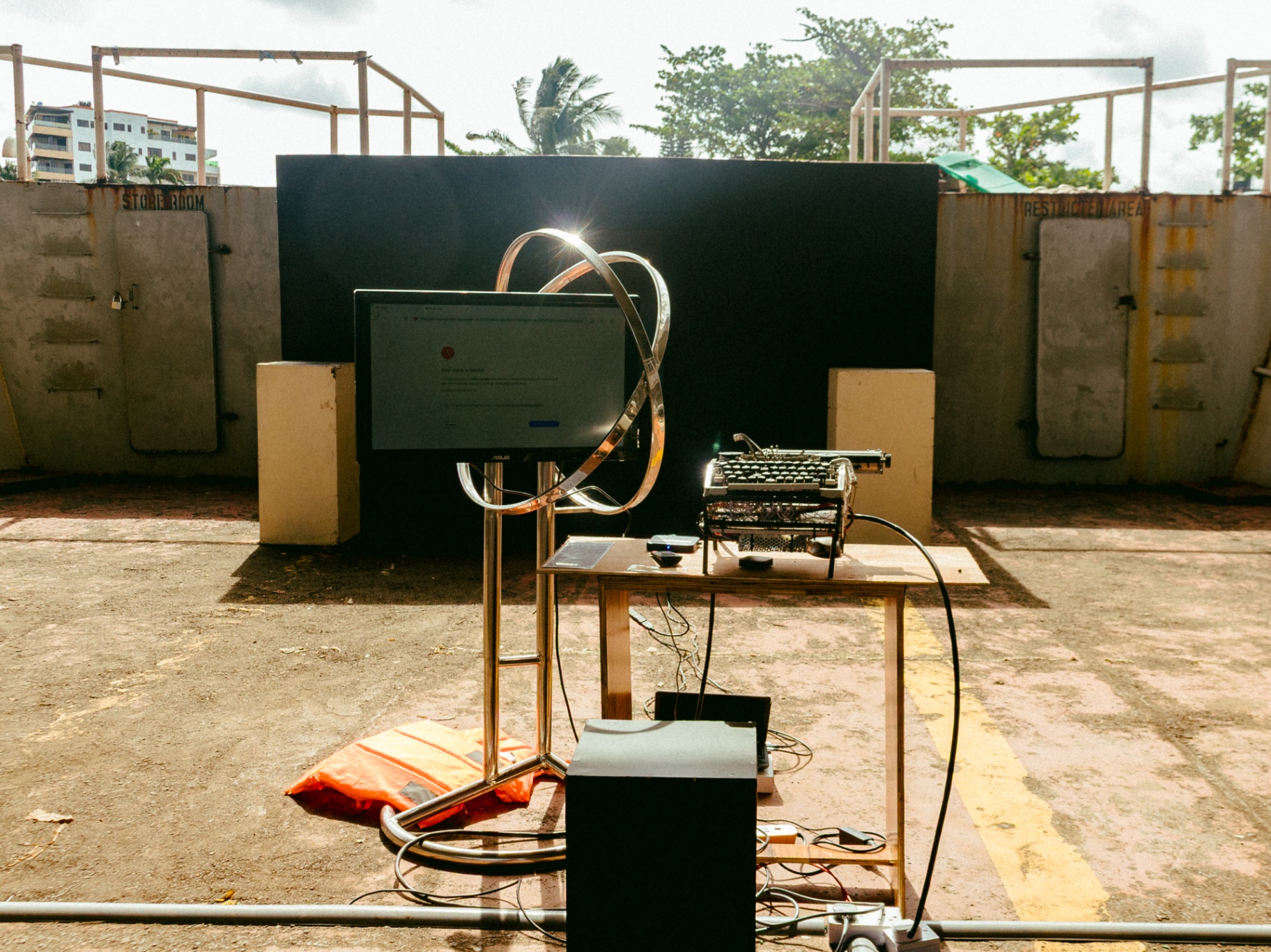 Outdoor installation with a monitor, chrome ring, and vintage typewriter on a wooden table against a black backdrop marked 'Store Room' and 'Restricted Area'
